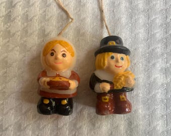 May include: Two small decorative candles in the shape of pilgrims. One is a woman holding a pie, the other a man holding wheat. Both are wearing traditional clothing and hats. Each candle has a string for hanging, ideal for seasonal decor.
