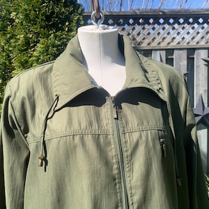 May include: An olive green jacket with a full zip, drawstring collar, and two zipped pockets. The jacket is displayed on a mannequin, highlighting its design. The colour and style suggest a casual, outdoor theme. The jacket is made of a lightweight material.