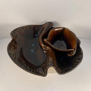 May include: A brown ceramic serving dish with a leaf-like design. The dish has a large, shallow section and a smaller, deeper section, connected by a curved element. The edges are decorated with a repeating pattern.