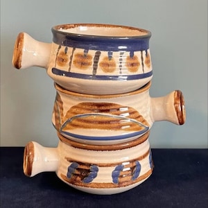 May include: Two stacked ceramic bowls with handles, in a cream colour with brown and blue painted patterns. Each bowl's rim has a dark blue band. These handmade bowls are suitable for serving soups or stews, and are likely of a rustic design.