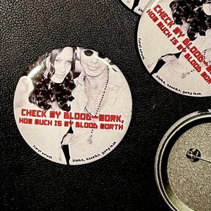 Może przedstawiać: Kolekcja okrągłych przypinek z czarno-białym zdjęciem dwóch osób. Przypinki mają czerwony tekst "CHECK MY BLOOD-WORK, HOW MUCH IS MY BLOOD WORTH". Przypinki leżą na czarnej, teksturowanej powierzchni.