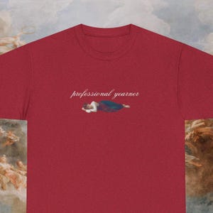 May include: A crimson t-shirt with the words "professional yearner" in cursive script. Beneath the text is a small illustration of a person reclining. The tee has a round neck and short sleeves.