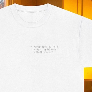 May include: White crew neck t-shirt with the handwritten text: "IF YOU'RE READING THIS I LIKED EVERYTHING BEFORE YOU DID." The shirt has a simple, casual design, suitable for everyday wear. The text is in a light grey colour.