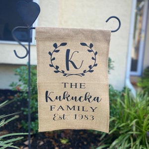 May include: A burlap garden flag with a black letter K in a wreath of leaves. The text "The Kukucka Family Est. 1983" is printed below the wreath.