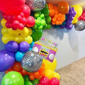 May include: A rainbow balloon arch with disco ball balloons and a pink and yellow boombox balloon. The arch is made of various colors of balloons, including red, orange, yellow, green, blue, purple, and pink.