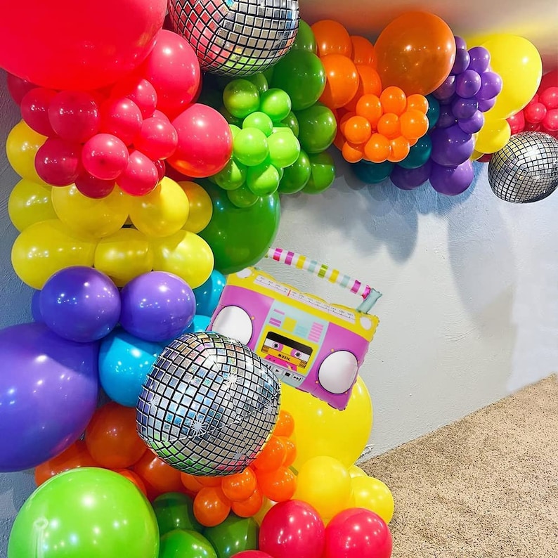 May include: A rainbow balloon arch with disco ball balloons and a pink and yellow boombox balloon. The arch is made of various colors of balloons, including red, orange, yellow, green, blue, purple, and pink.