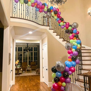 May include: A staircase decorated with a balloon garland. The garland is made of colorful balloons, including pink, green, blue, orange, purple, and black. There are also silver disco ball balloons in the garland.