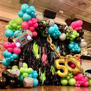 May include: A black backdrop with neon green and pink streamers, a gold chain, and a large gold number 50. The backdrop is decorated with a variety of balloons in blue, green, pink, and black. Some of the balloons are shaped like disco balls and a boombox.