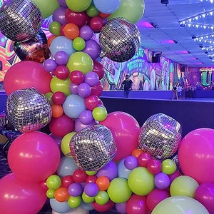 May include: A colorful balloon arch with disco ball balloons, pink, green, purple, and blue balloons. The arch is set up in a roller skating rink with a black and white checkered floor.