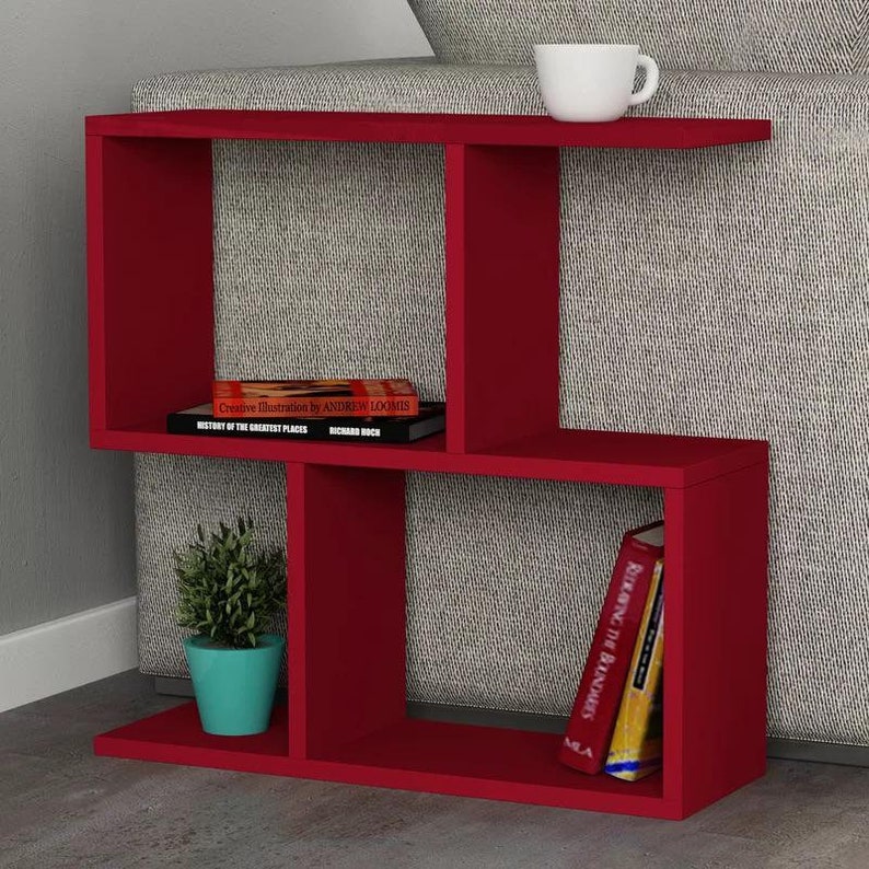 May include: Red wooden side table with three shelves. The table has a unique design with a shelf that extends out from the side. The table is decorated with a potted plant, a cup, and two books. The books are titled "Creative Illustration by Andrew Loomis" and "Remaking the Boundaries".