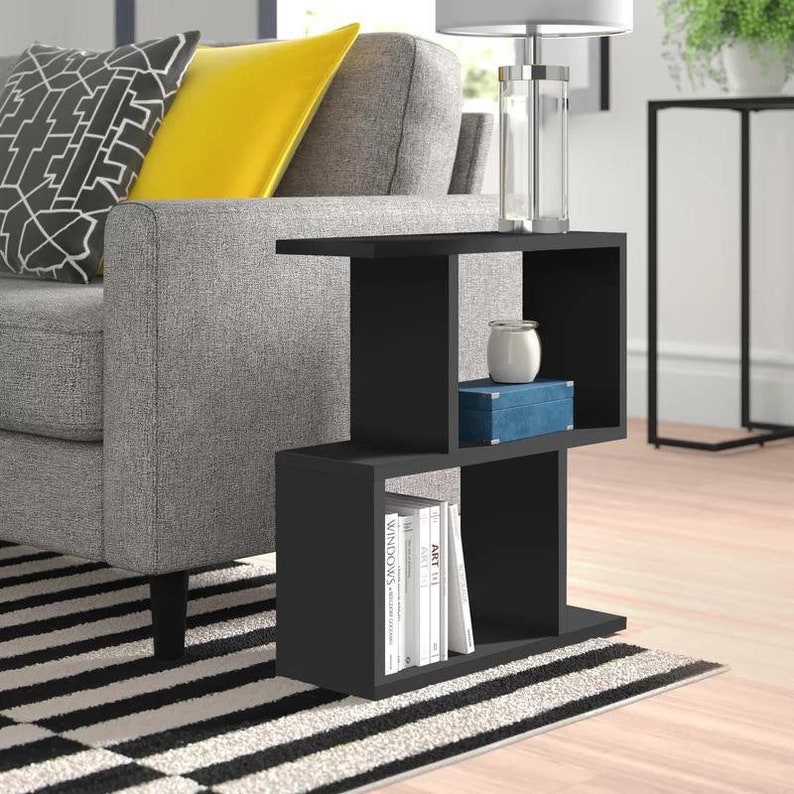 May include: Black side table with three shelves. The top shelf has a clear glass lamp with a silver base. The middle shelf has a blue box and a white candle. The bottom shelf has a stack of books with titles like "Art" and "Windows".