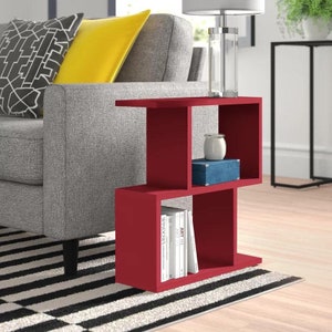 May include: A red end table with three shelves. The top shelf has a white ceramic vase and a blue box. The bottom shelf has a stack of books with titles like "Art" and "Windows".