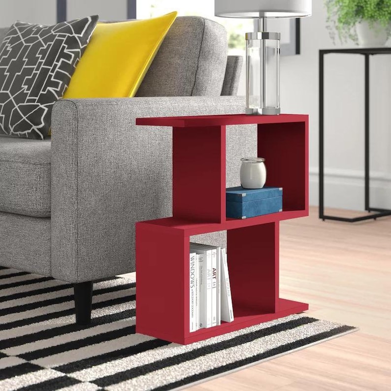 May include: A red end table with three shelves. The top shelf has a white ceramic vase and a blue box. The bottom shelf has a stack of books with titles like "Art" and "Windows".