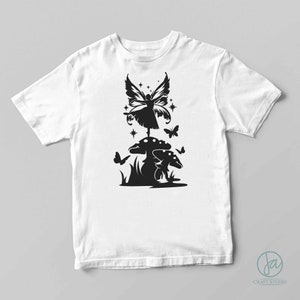 Puede incluir: Camiseta blanca con un dise&ntilde;o negro de una hada sobre una seta, rodeada de mariposas y estrellas. El dise&ntilde;o presenta siluetas detalladas del hada, las setas y el follaje, creando una est&eacute;tica caprichosa y encantadora.