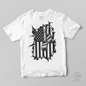 May include: White t-shirt featuring a black graphic of a torn American flag with dog tags. The flag design includes stars and stripes, with two dog tags hanging from a chain. The shirt is a classic crew neck style.