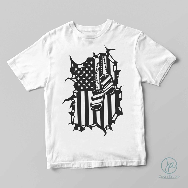 May include: White t-shirt featuring a black graphic of a torn American flag with dog tags. The flag design includes stars and stripes, with two dog tags hanging from a chain. The shirt is a classic crew neck style.