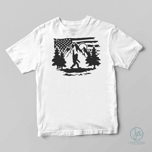 May include: A white t-shirt featuring a black silhouette of Bigfoot standing in front of a mountain range with a full moon and an American flag in the background.