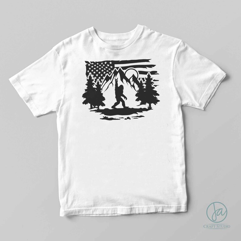May include: A white t-shirt featuring a black silhouette of Bigfoot standing in front of a mountain range with a full moon and an American flag in the background.