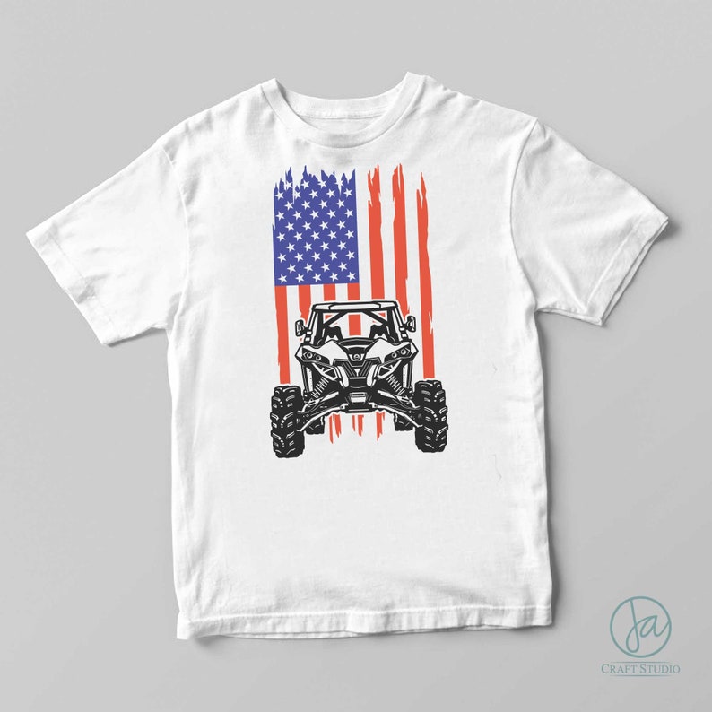 Pode incluir: Camiseta branca com um gr&aacute;fico de um ve&iacute;culo off-road preto, em um design de bandeira americana desgastada em vermelho, branco e azul. A camiseta tem mangas curtas e gola redonda.