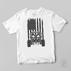 Pode incluir: Camiseta branca com um gr&aacute;fico preto de um ve&iacute;culo off-road e um design de bandeira americana. A bandeira tem listras verticais desgastadas e um campo de estrelas. O design est&aacute; centrado na frente da camiseta.