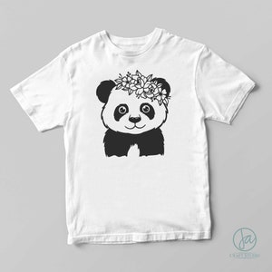 May include: A white t-shirt with a black and white panda bear wearing a flower crown. The panda is smiling and looking at the viewer.
