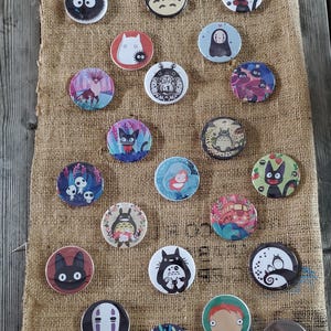 Peut inclure: Une collection de vingt-deux badges ronds épinglés présentant diverses illustrations de personnages de dessins animés colorés, incluant des créatures aux grands yeux, des esprits de la forêt et des figures disposées sur un tissu marron texturé.