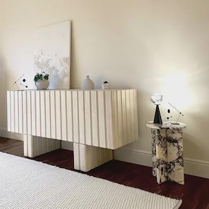 May include: A contemporary room setting with a large, light-toned wooden cabinet with vertical slats. A white plinth, a small side table with a marble pattern, and a white rug are also present. The room has a minimalist design with neutral colours.