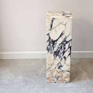 May include: A white marble pedestal with brown and black veining. The pedestal is square and has a smooth, polished surface.