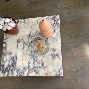 Op de afbeelding: Een vierkante marmeren salontafel met een bruine vaas gevuld met witte wattenbolletjes, een glazen kom met een bruine steen en een terracotta vaas erop.