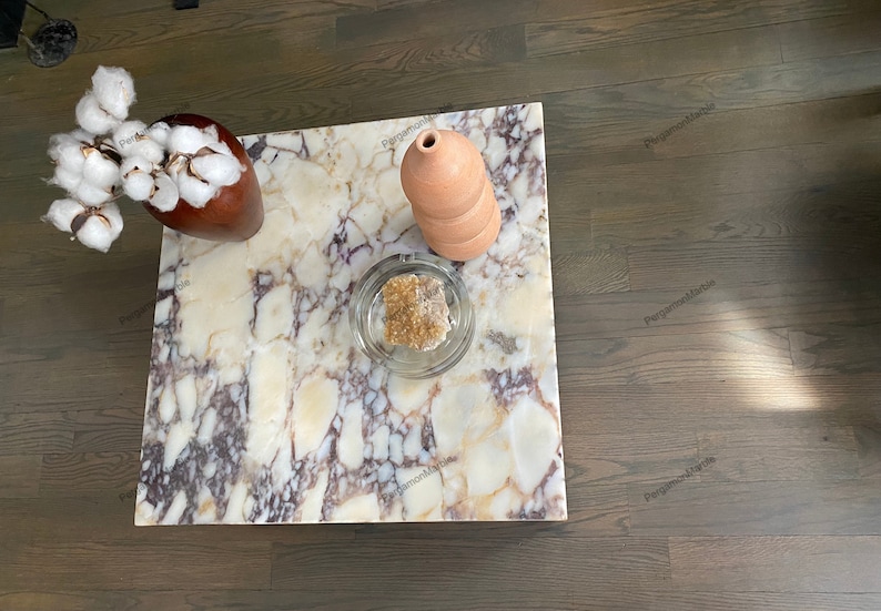 Op de afbeelding: Een vierkante marmeren salontafel met een bruine vaas gevuld met witte wattenbolletjes, een glazen kom met een bruine steen en een terracotta vaas erop.