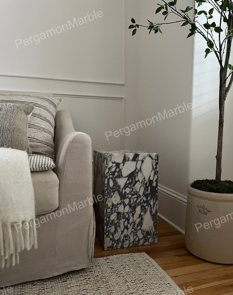 K&ouml;nnte beinhalten: Ein schwarzer und wei&szlig;er Marmor-Beistelltisch mit einer quadratischen Oberseite und einem rechteckigen Sockel. Der Tisch steht auf einem Holzboden in einem Raum mit einer wei&szlig;en Wand und einem Sofa.