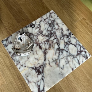 Op de afbeelding: Een wit marmeren dienblad met paarse en bruine aderen. Het dienblad ligt op een houten oppervlak en heeft een zilveren kom erop.