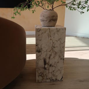 May include: A white marble pedestal with a brown and white ceramic vase with green leafy branches on top. The pedestal is sitting on a wooden floor.