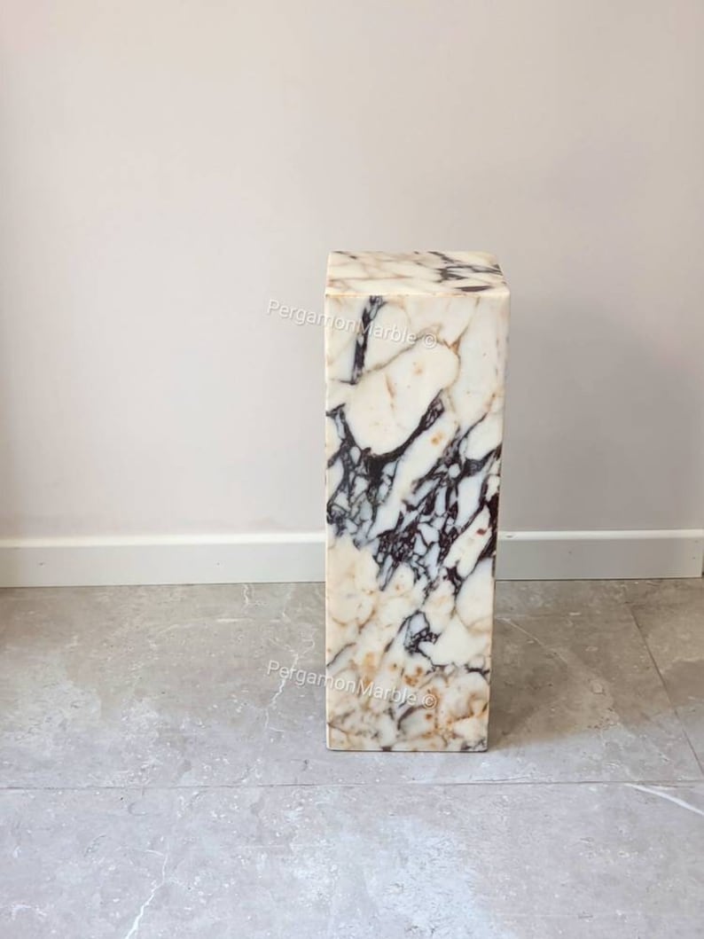 May include: A white marble pedestal with brown and black veining. The pedestal is square and has a smooth, polished surface.