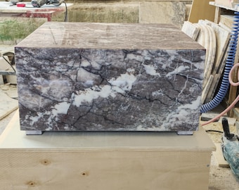 May include: A rectangular block made of polished dark purple and white marble with prominent veining throughout the surface. The stone object features sharp, clean edges and sits on small, square, light-colored feet at each of the four bottom corners.