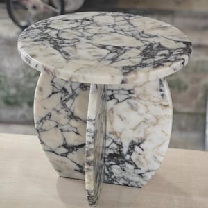 May include: A round marble side table with a white and gray veined pattern. The table has a circular top and a base with three curved supports. The table is approximately 18 inches in diameter and 18 inches tall.
