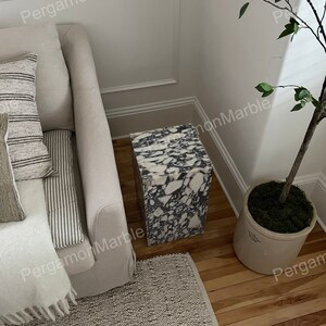 K&ouml;nnte beinhalten: Ein wei&szlig;er und schwarzer Marmor-Beistelltisch mit quadratischer Tischplatte und einem wei&szlig;en und grauen Teppich darunter. Der Tisch steht in einer Ecke eines Raumes mit einem Sofa und einer Topfpflanze.