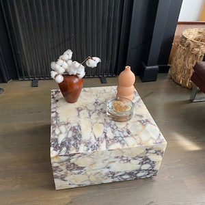 Op de afbeelding: Een vierkante, witte marmeren salontafel met bruine en gouden aderen. De tafel staat op een houten vloer. Een kleine vaas met witte katoenen bloemen en een terracotta vaas staan op de tafel.