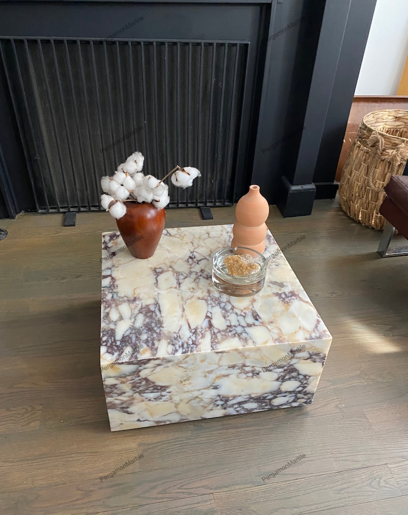 Op de afbeelding: Een vierkante, witte marmeren salontafel met bruine en gouden aderen. De tafel staat op een houten vloer. Een kleine vaas met witte katoenen bloemen en een terracotta vaas staan op de tafel.