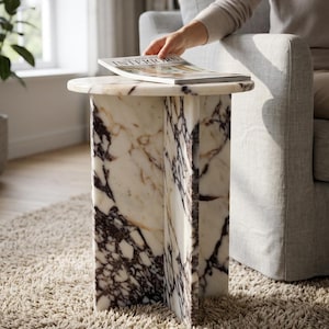 May include: A round side table crafted from white marble with prominent dark purple and black veining. The table features a cylindrical top supported by a cross-shaped base made of two intersecting vertical marble slabs, showcasing a natural stone pattern.