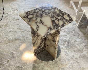 May include: A square marble side table with a white, purple, and gold marbled top and a white marble base. The table is sitting on a concrete floor.