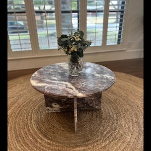 May include: A round, pink and white marble coffee table with a natural jute rug underneath. A vase with white flowers sits on the table.