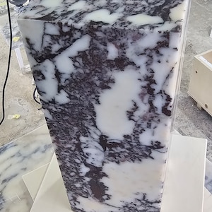 May include: A large, white marble block with purple and black veining. The block is sitting on a white surface.