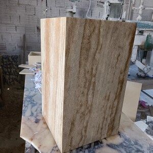 May include: A rectangular, beige stone block with vertical brown veining. The block is in a workshop setting, resting on a marble surface. The stone has a natural, textured appearance, showcasing its unique grain patterns. The block is a solid, geometric shape.