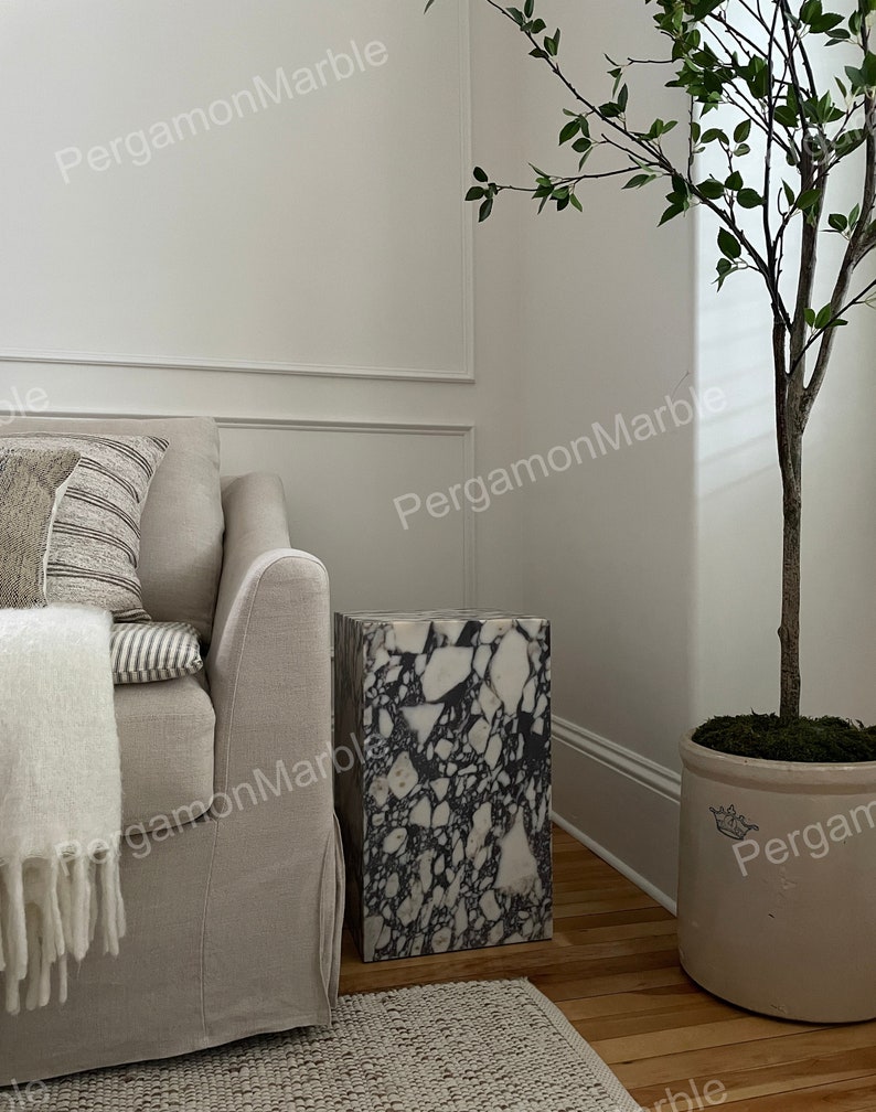K&ouml;nnte beinhalten: Ein wei&szlig;er und schwarzer Terrazzo-Beistelltisch mit einem wei&szlig;en Sofa und einer Topfpflanze im Hintergrund.