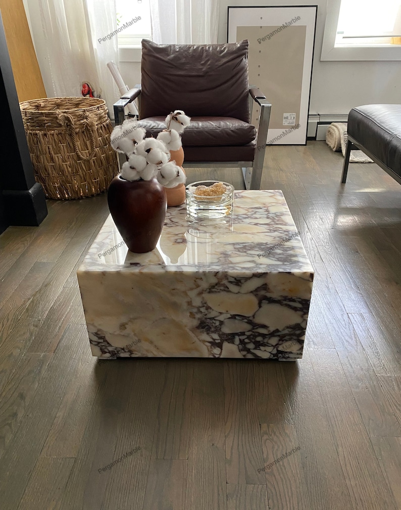 Op de afbeelding: Een vierkante marmeren salontafel met een wit, bruin en paars marmerpatroon. Een bruine vaas met witte katoenen bloemen staat op de tafel, samen met een glazen kom.