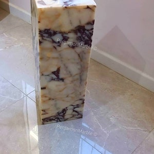 May include: A white marble pedestal with brown and purple veining. The pedestal is square and has a smooth, polished surface. The text "PergamonMarble &copy;" is visible on the side of the pedestal.