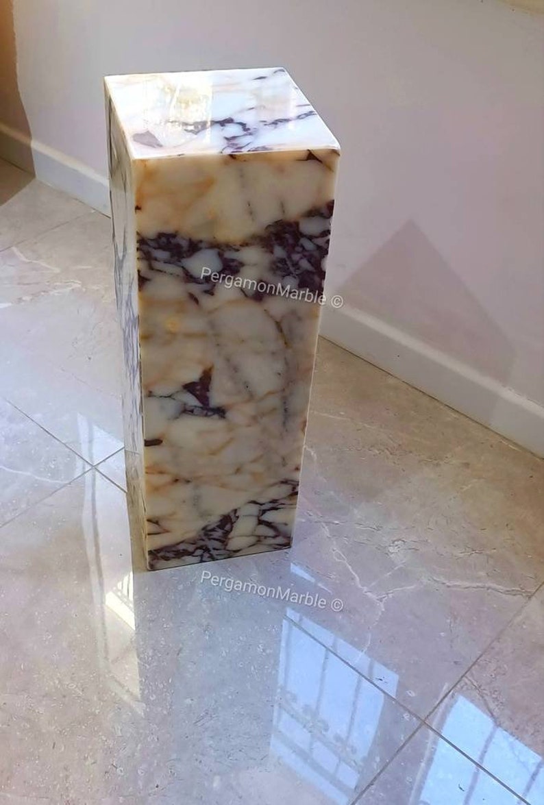May include: A white marble pedestal with brown and purple veining. The pedestal is square and has a smooth, polished surface. The text "PergamonMarble &copy;" is visible on the side of the pedestal.