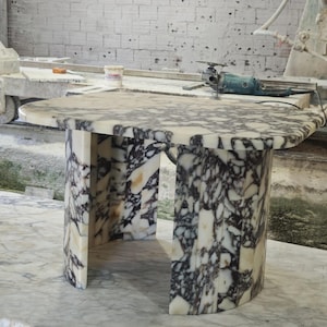 May include: A marble table with a rounded top and a unique base design. The table features a white and black patterned surface. The base has a curved shape with an open center. The table is approximately 76 cm long.
