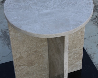 May include: A round marble side table with a light beige color and a veined pattern. The table has a circular top and a base with four rectangular supports. The table is approximately 20 inches in diameter.